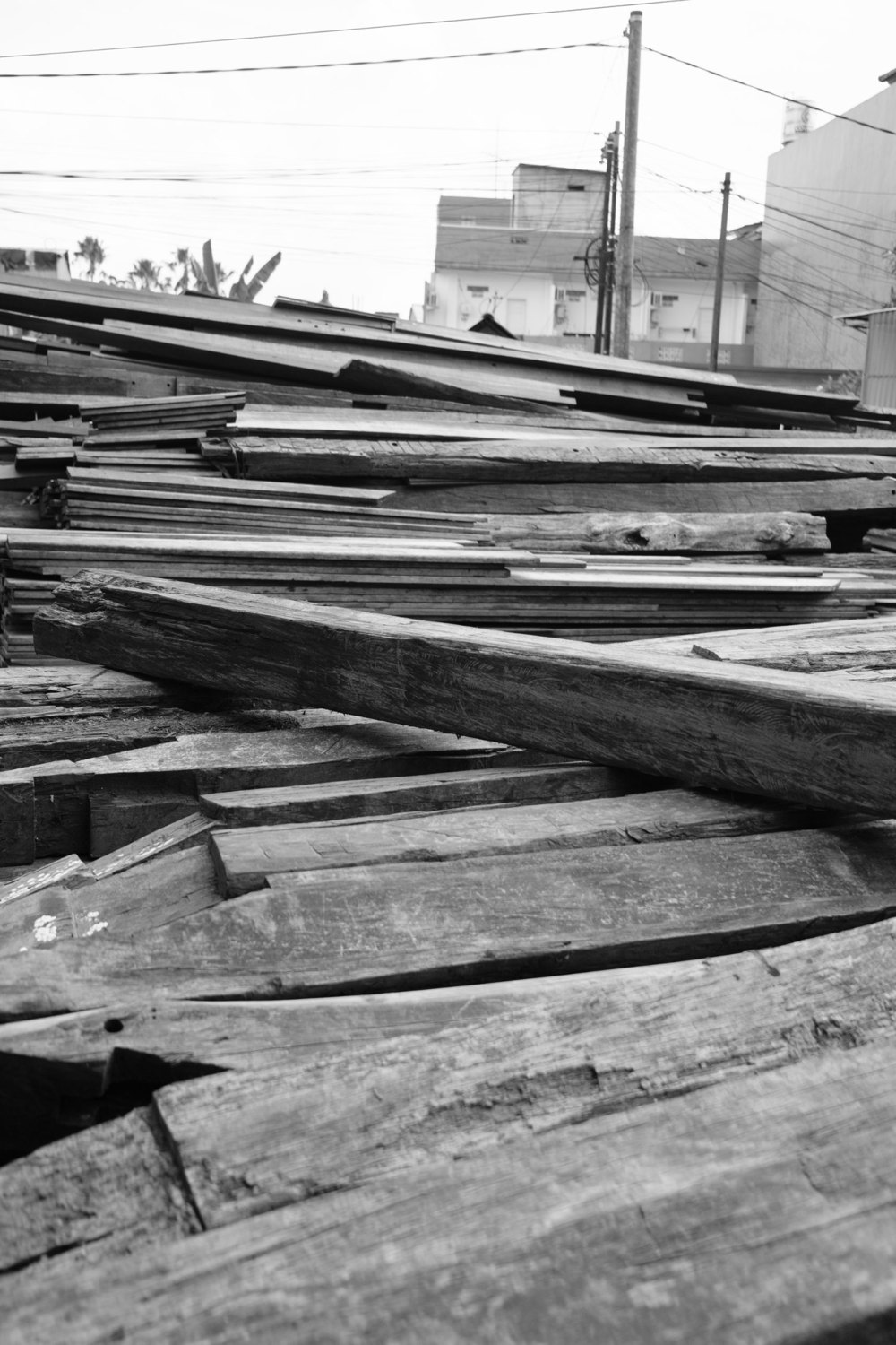 Black-and-white photograph of old boat planks and beams piled in a timber yard, houses and power lines behind