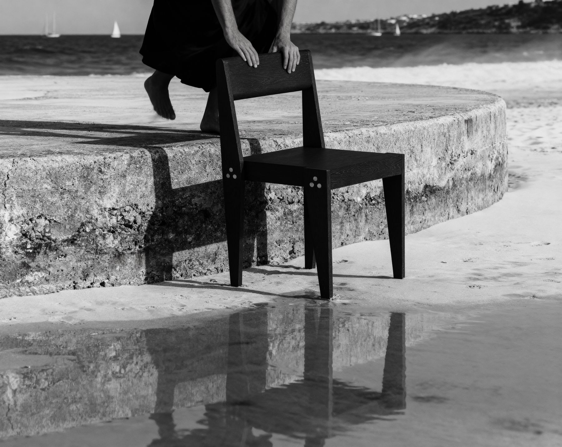 Black-and-white photograph of hands resting on the backrest of The Chair on a beach, waves and sailboats behind