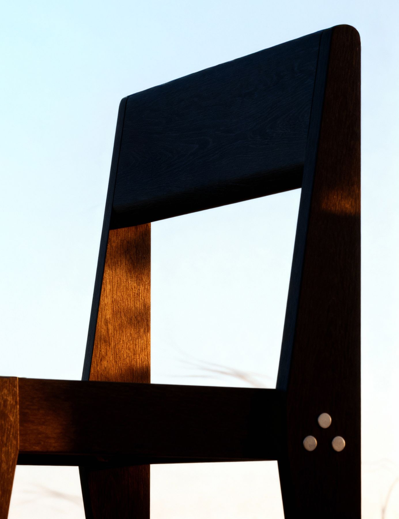 The Chair's backrest against an evening sky, with low sun on the teak and the bolt cluster on the rear leg