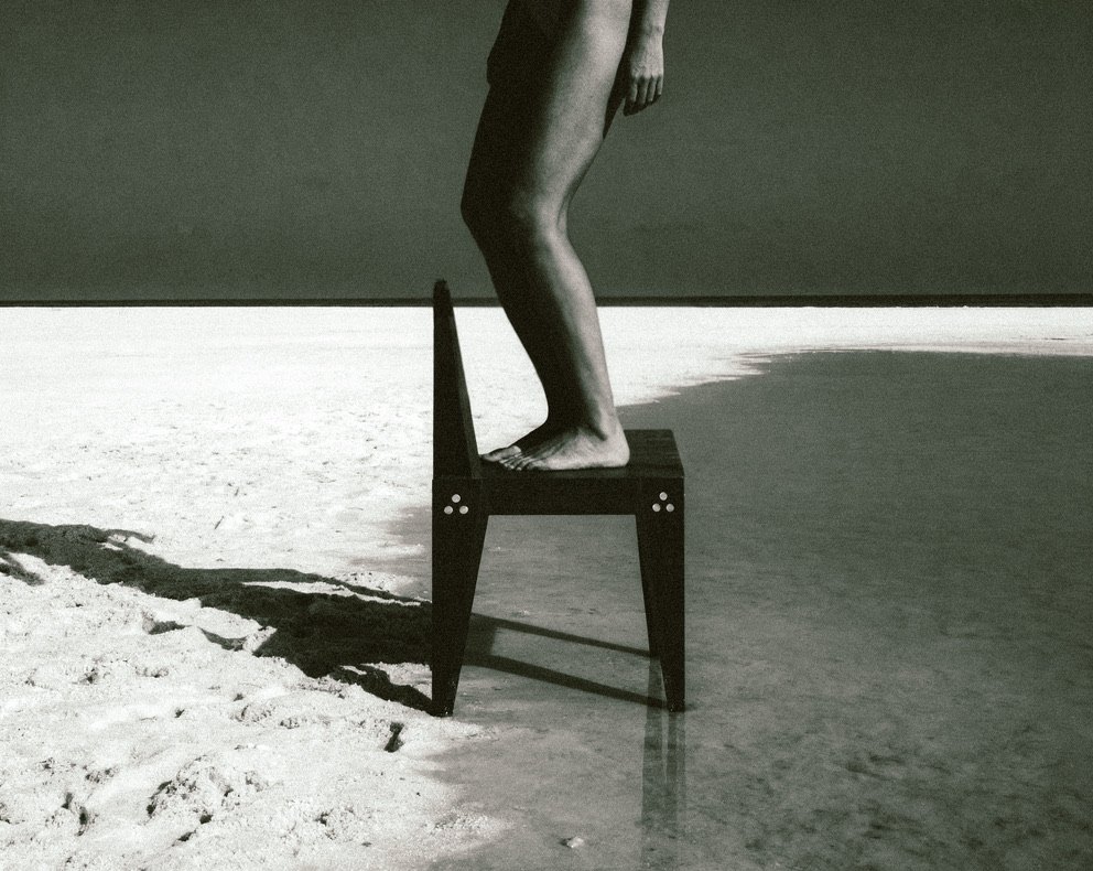 Black-and-white photograph of bare legs standing on The Chair at the edge of shallow water on a salt flat